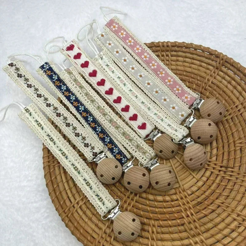 A collection of fabric pacifier chains with flower and heart patterns and wooden clips on a woven tray.