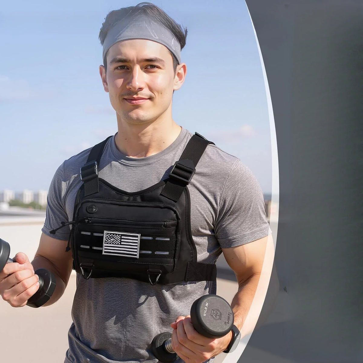 A man in a grey shirt holding dumbbells while wearing a black tactical chest bag.