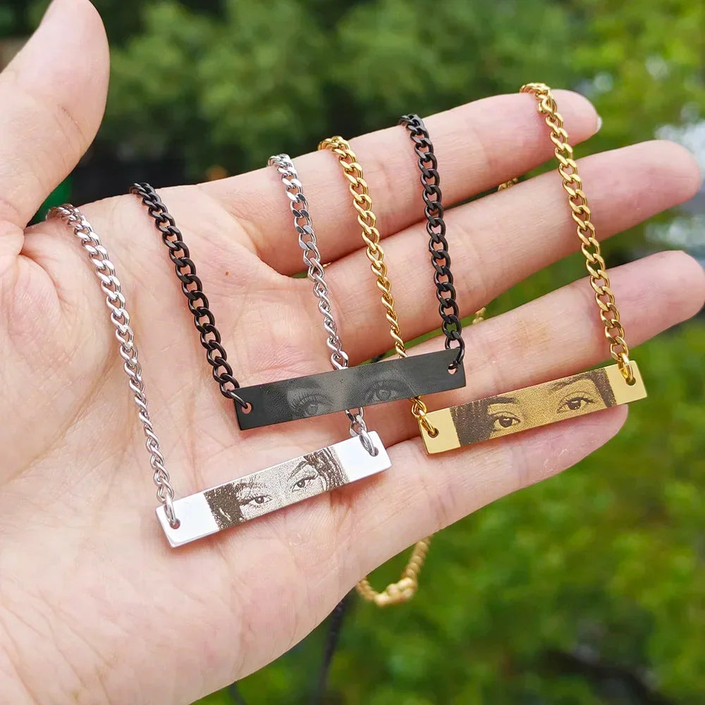 Hand holding three personalized photo engraved bar necklaces in silver, black, and gold tones with heart cutouts.