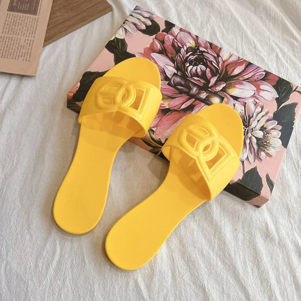 Pair of yellow jelly slides with a decorative cutout letter emblem on the strap, resting on a floral box.