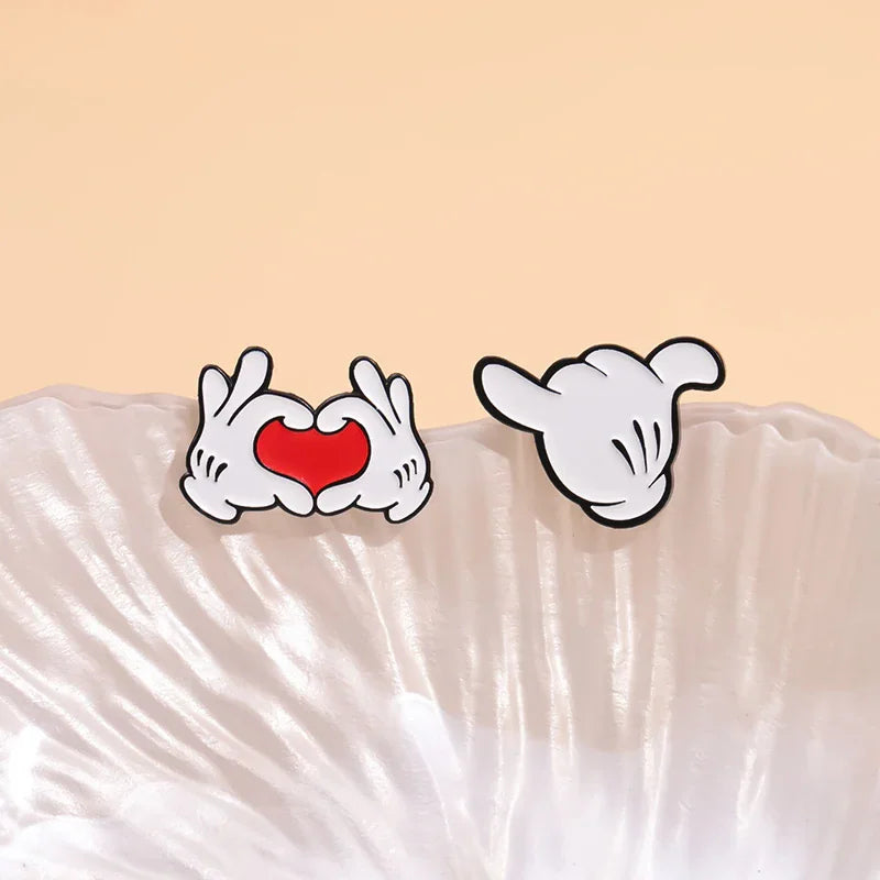 Two cartoon hand gesture enamel pins showing a heart and a shaka sign placed on a white seashell dish