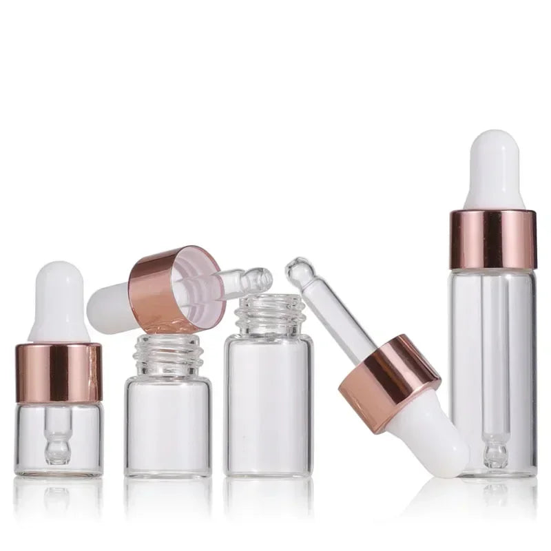 A group of clear mini glass dropper bottles with rose gold caps and white silicone droppers, displayed upright and on their