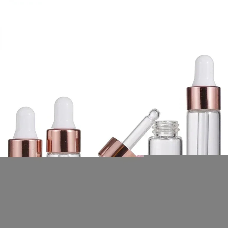 Several clear mini glass dropper bottles with rose gold caps and white silicone droppers, some lying on their sides.