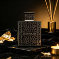 Black rectangular perfume bottle with intricate geometric gold-toned pattern and matching cap on a marble base with candle