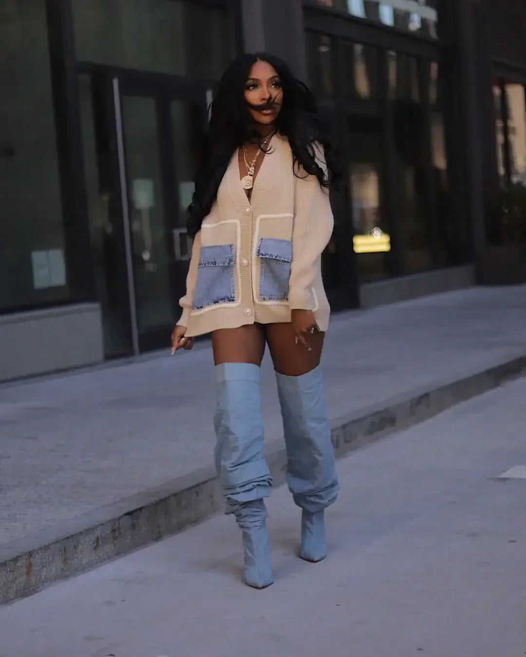 A woman wearing a beige knit cardigan with blue denim pockets and slouchy denim over-the-knee boots.