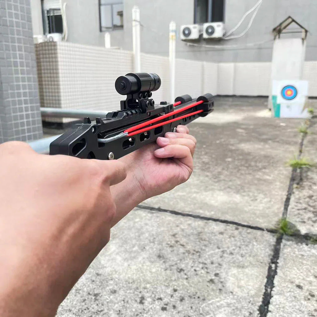 Person aiming a black hunting slingshot at a paper target on a balcony