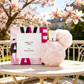 A pale pink perfume bottle with a fluffy pom-pom and its box placed on a wooden table outdoors with blooming magnolia trees.