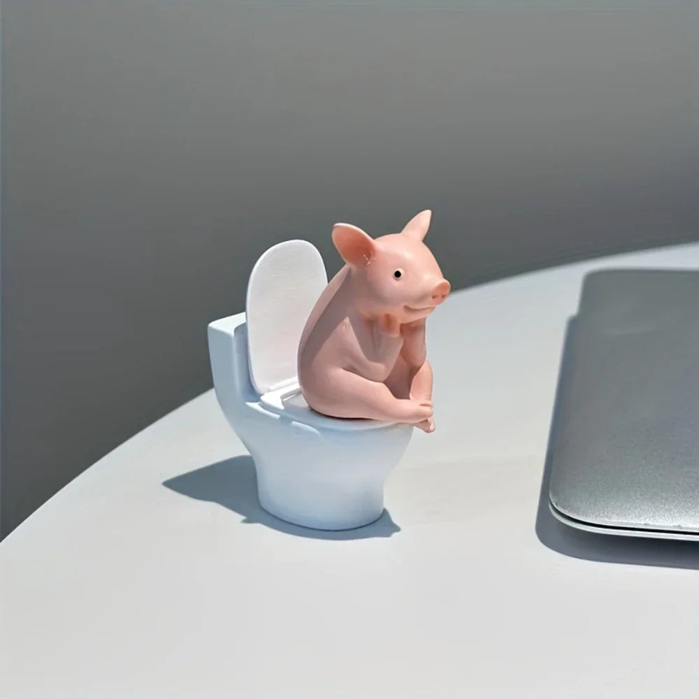 A cute pink pig figurine sitting on a miniature white toilet, displayed on a desk.