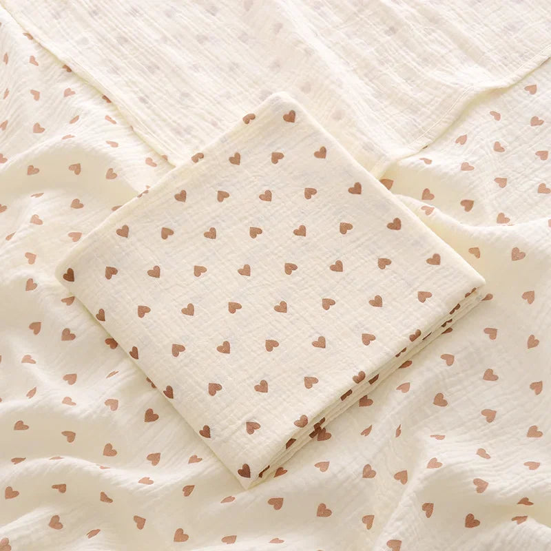 Cream-colored cotton muslin baby swaddle blanket with a brown heart pattern, folded.