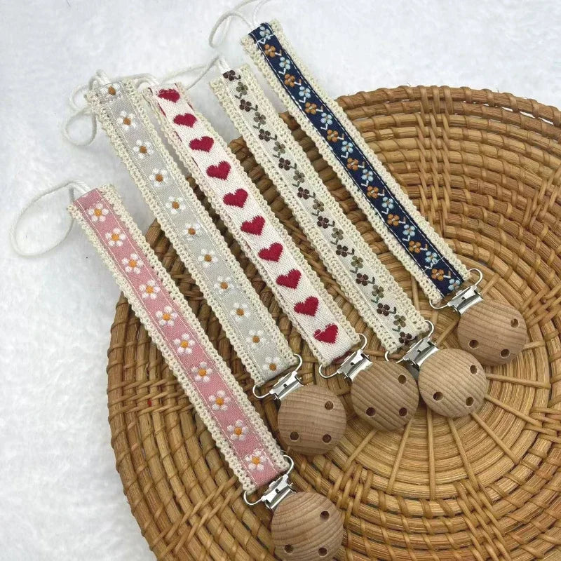 Several fabric pacifier chains with floral and heart patterns and wooden clips on a woven tray.
