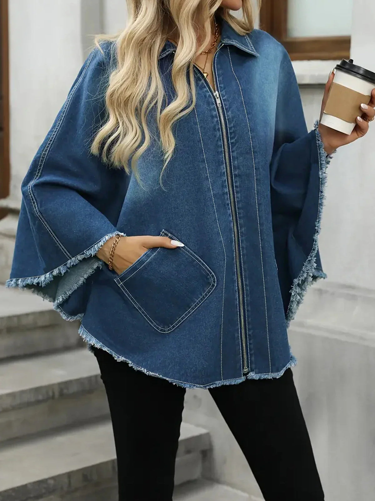 A close-up view of a blue denim jacket with a frayed hem, worn by a woman holding a coffee cup