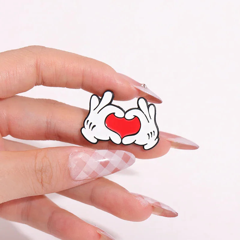 A hand holding a cartoon hand gesture enamel pin showing a heart shape