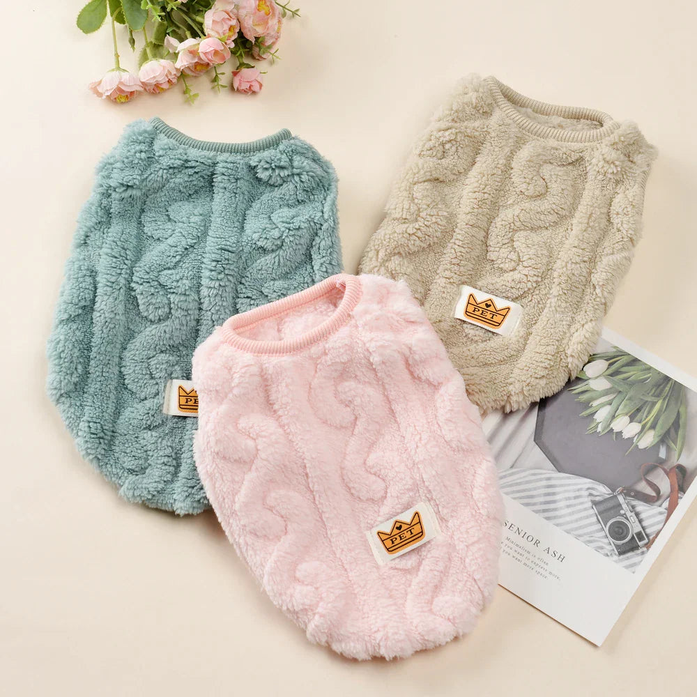 Three colors of soft fleece dog sweaters arranged with flowers on a flat surface.