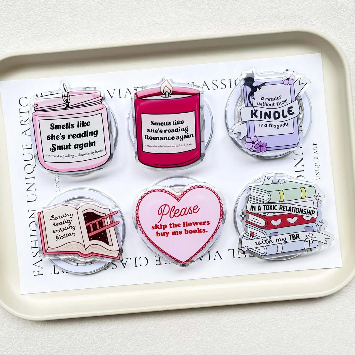 Assortment of six book-themed acrylic phone grips placed on a white serving tray.