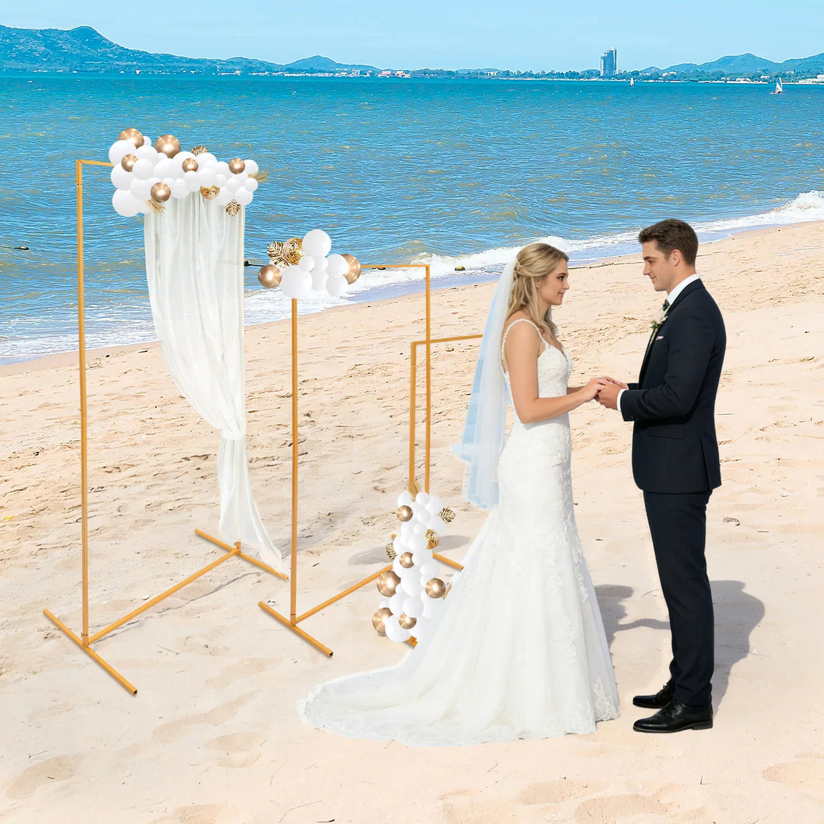 Gold metal wedding arch display stands decorated with balloons and fabric on a beach