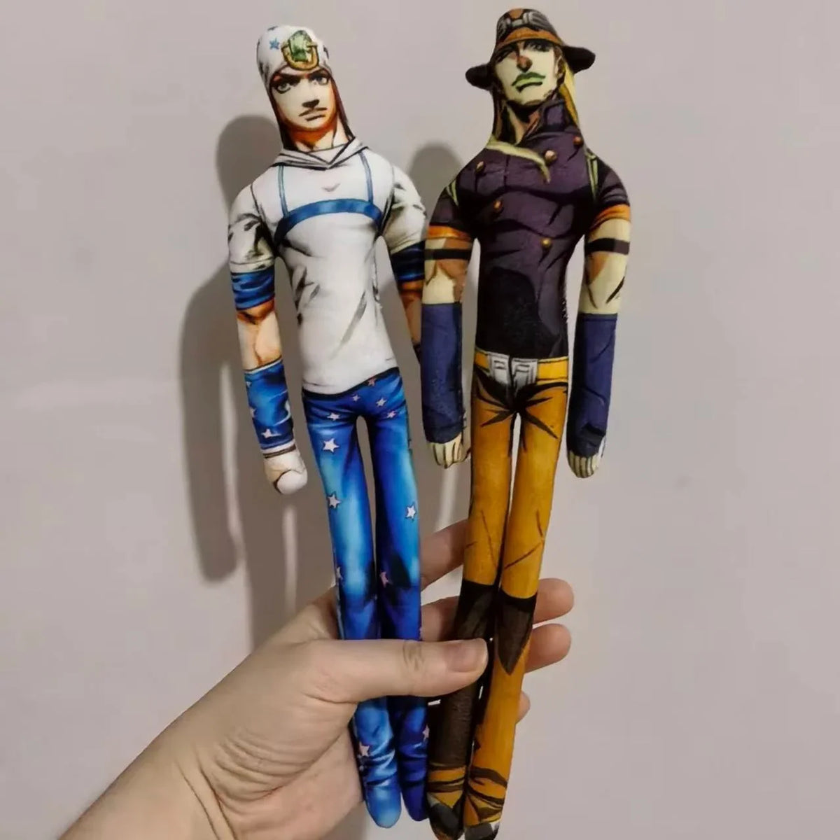 Hand holding two anime character plush dolls wearing hats