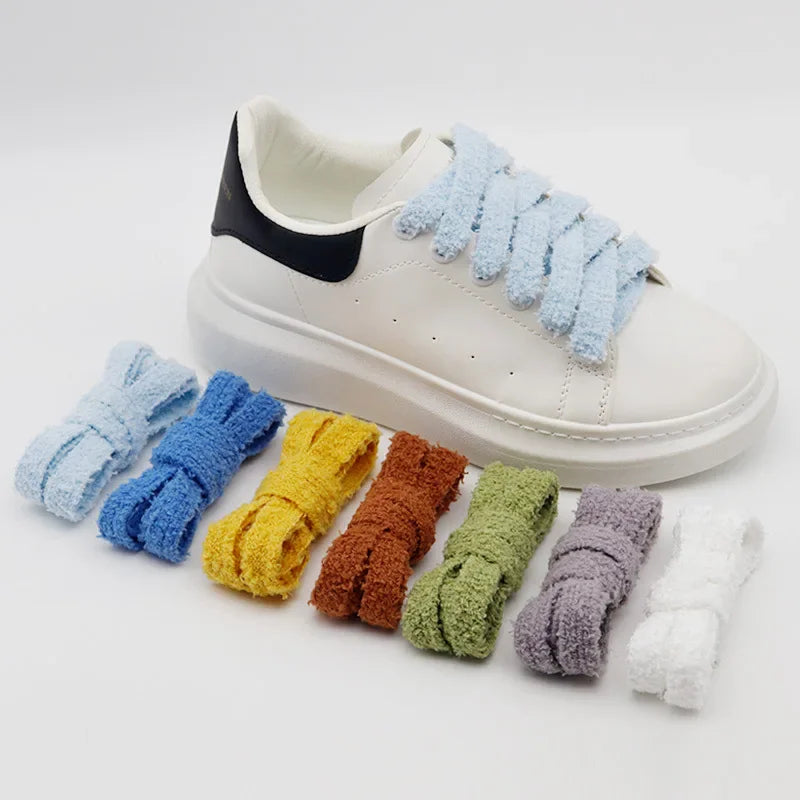 A white sneaker with blue fluffy shoelaces tied, surrounded by additional pairs of folded plush shoelaces in various colours.