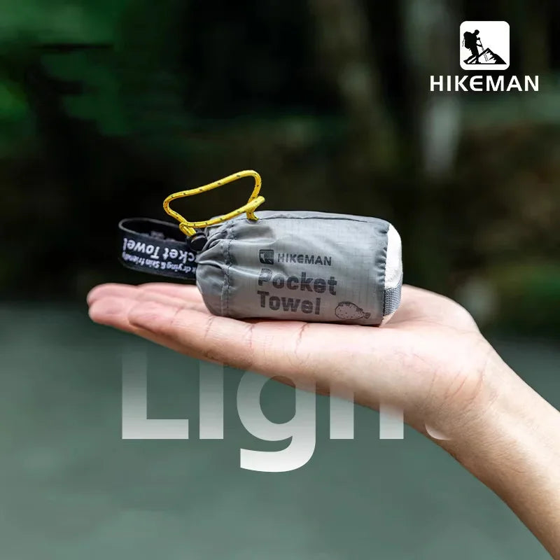 A hand holding a compact, folded grey pocket towel with a yellow carabiner in an outdoor setting.