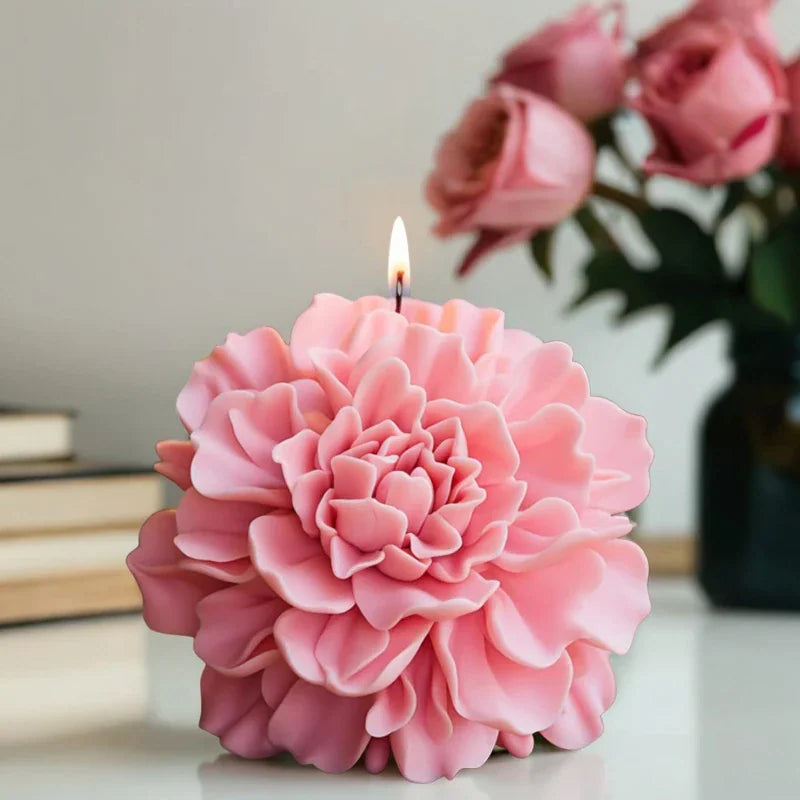 A single lit pink peony shaped flower candle on a table with a rose bouquet