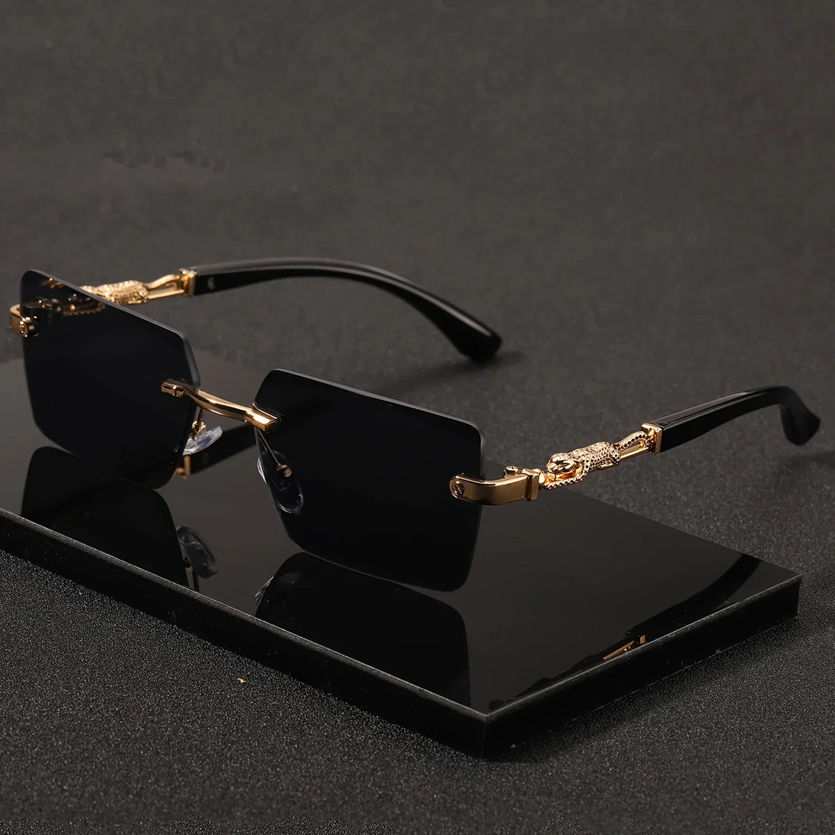 Rimless rectangular sunglasses with black lenses and gold panther-style metal temples on a black display base