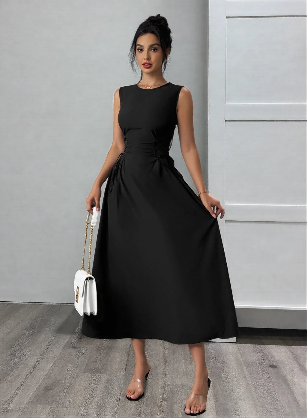 A woman wearing a black sleeveless midi dress with side ties, holding a white bag.