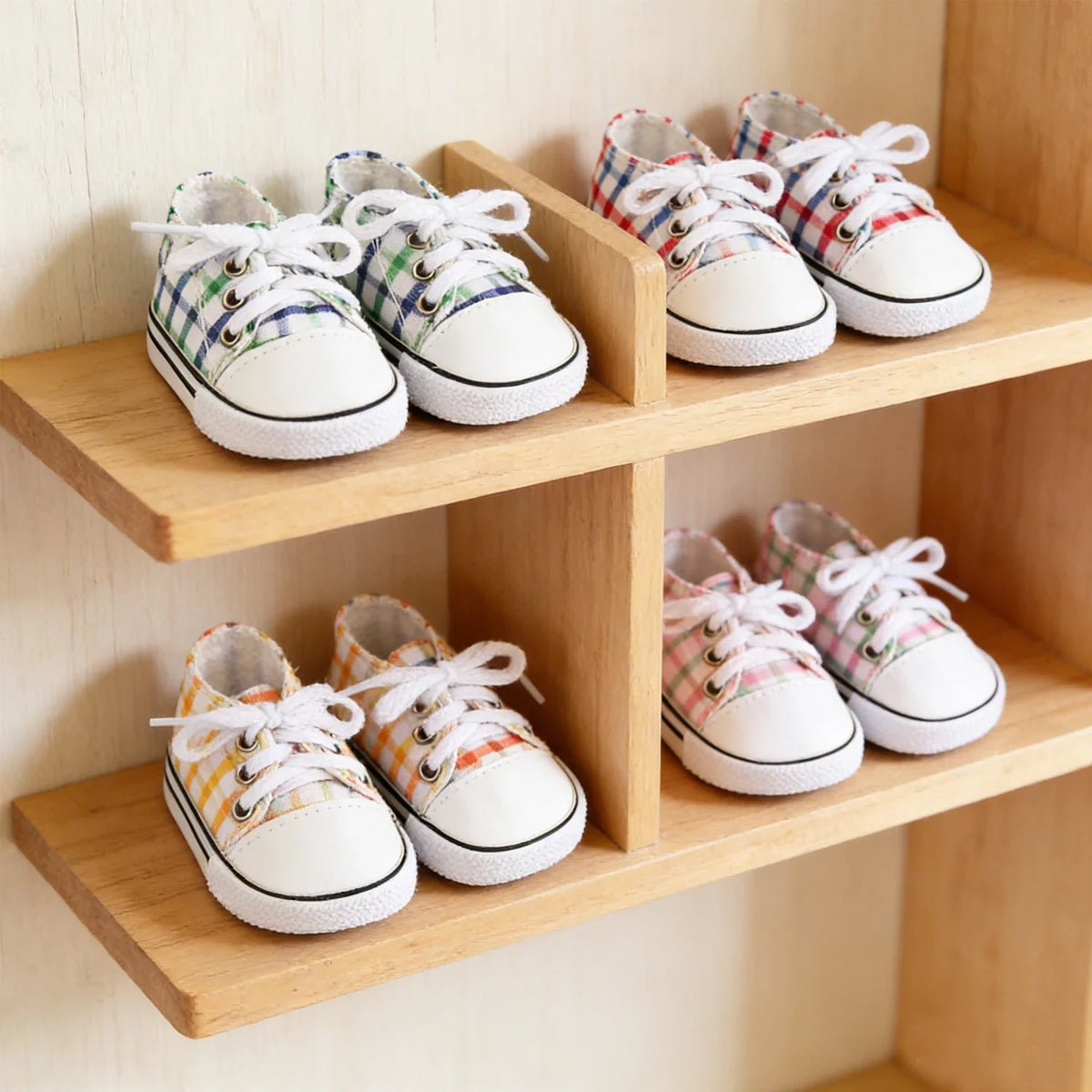 Four pairs of colorful checkered canvas doll shoes with white laces and toes, displayed on a wooden shelf.
