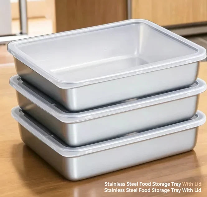 Stackable rectangular stainless steel food storage trays with clear lids on a wooden surface