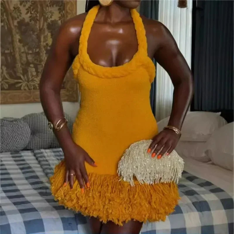 A woman wearing a mustard yellow velvet halter mini dress with braided straps and fringe hem, holding a beaded clutch.