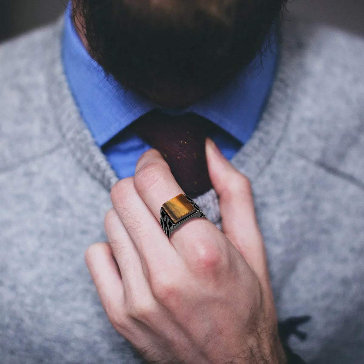 A man's hand wearing a square tiger eye signet ring, adjusting his tie