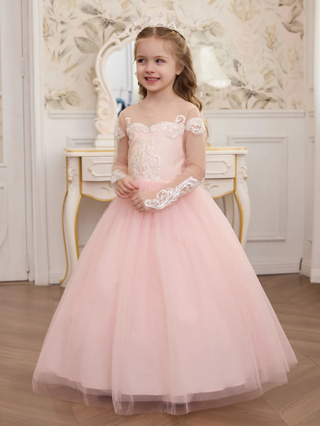 A young girl standing in a pale pink formal tulle dress with lace embroidery on the sheer sleeves and bodice.
