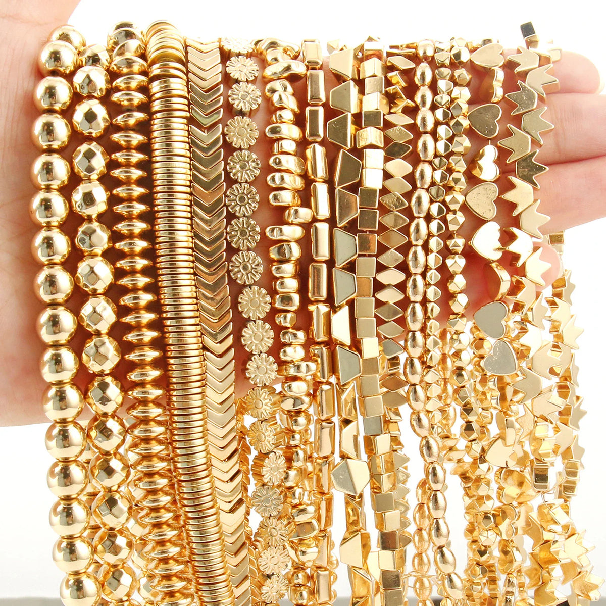 A hand holding various strands of gold-plated hematite beads in round, cube, heart, and spacer shapes.