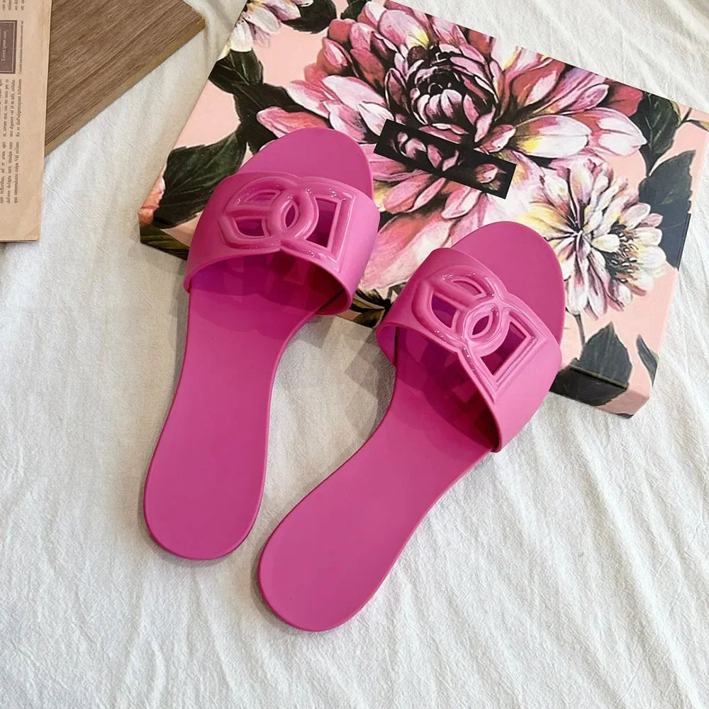 Pair of fuchsia jelly slides with a decorative cutout letter emblem on the strap, resting on a floral box.