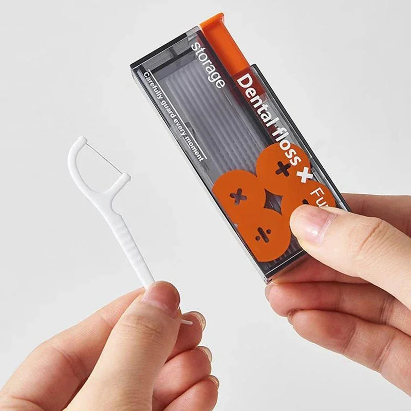 A person using their hand to extract a single white plastic dental floss stick from a grey and orange dispenser.