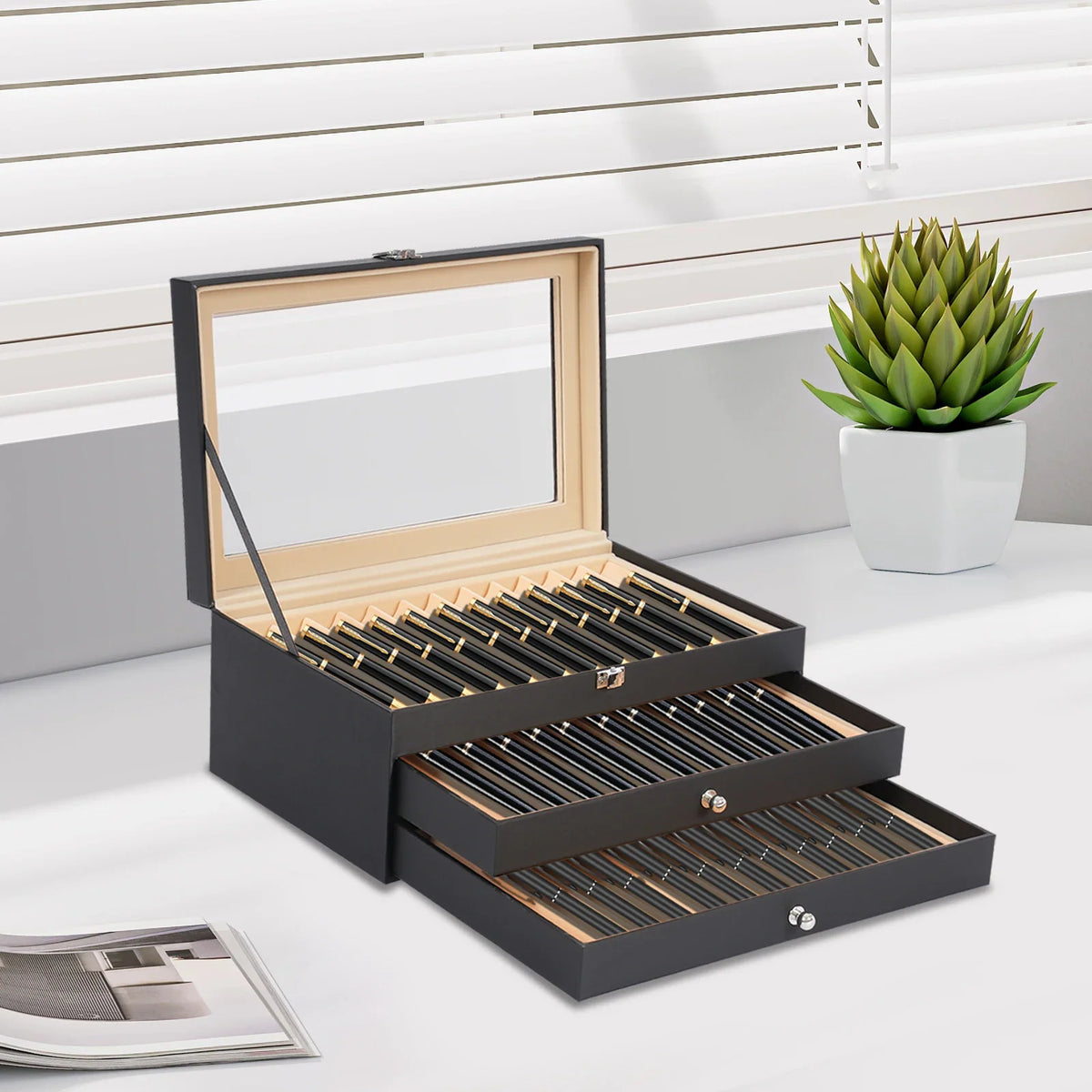 Open black 36-slot pen display case on a desk with a plant.