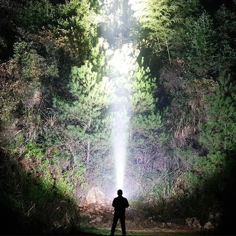 Person standing outdoors at night using a powerful handheld tactical flashlight to illuminate trees.