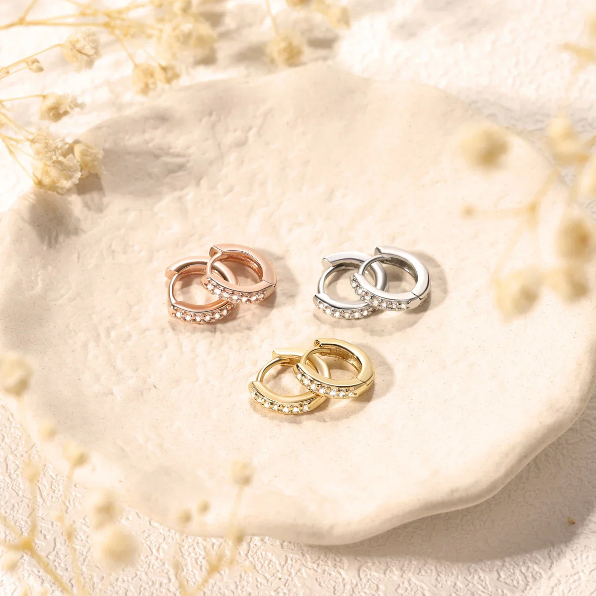 Three pairs of small crystal hoop earrings in silver, gold and rose gold placed on a small ceramic dish.