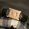 A rose gold rectangular dial watch with a green leather strap placed on a plaid patterned cloth.