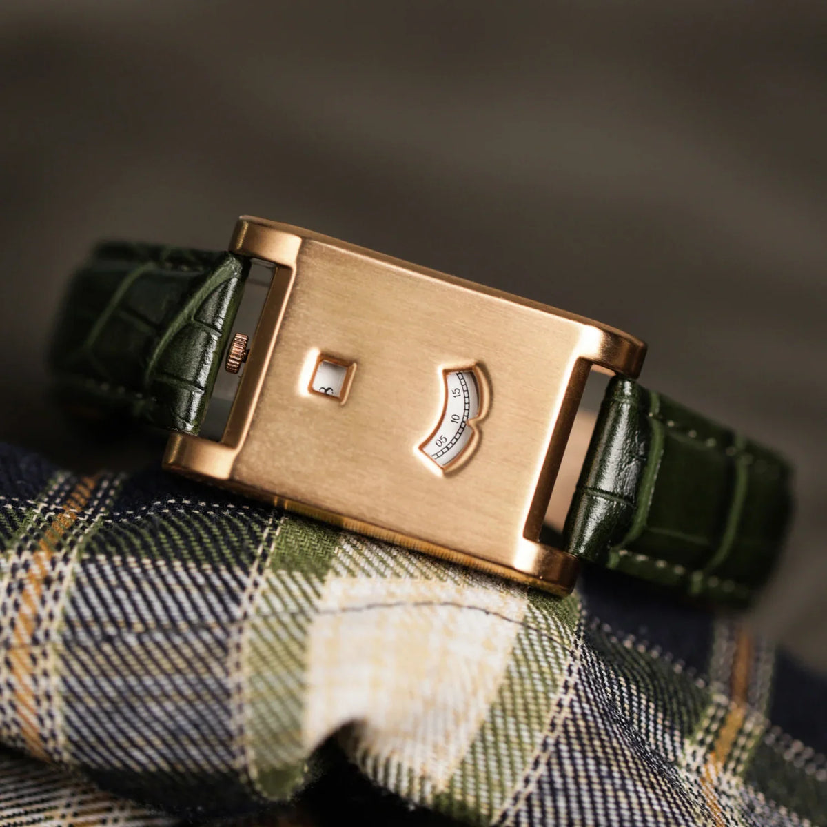 A rose gold rectangular dial watch with a green leather strap placed on a plaid patterned cloth.
