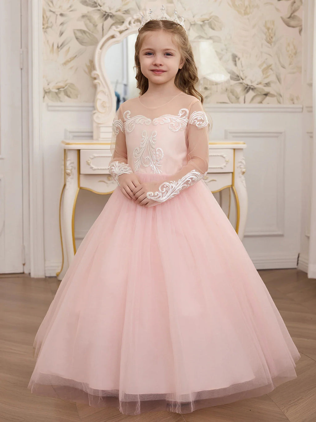 A young girl posing in a pale pink tulle formal dress with intricate white lace detailing on the bodice.