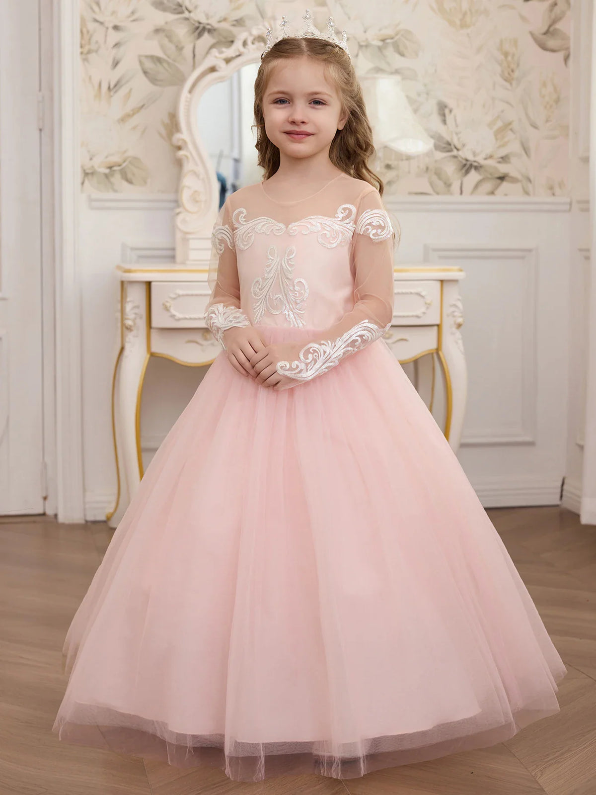 A young girl posing in a pale pink tulle formal dress with intricate white lace detailing on the bodice.
