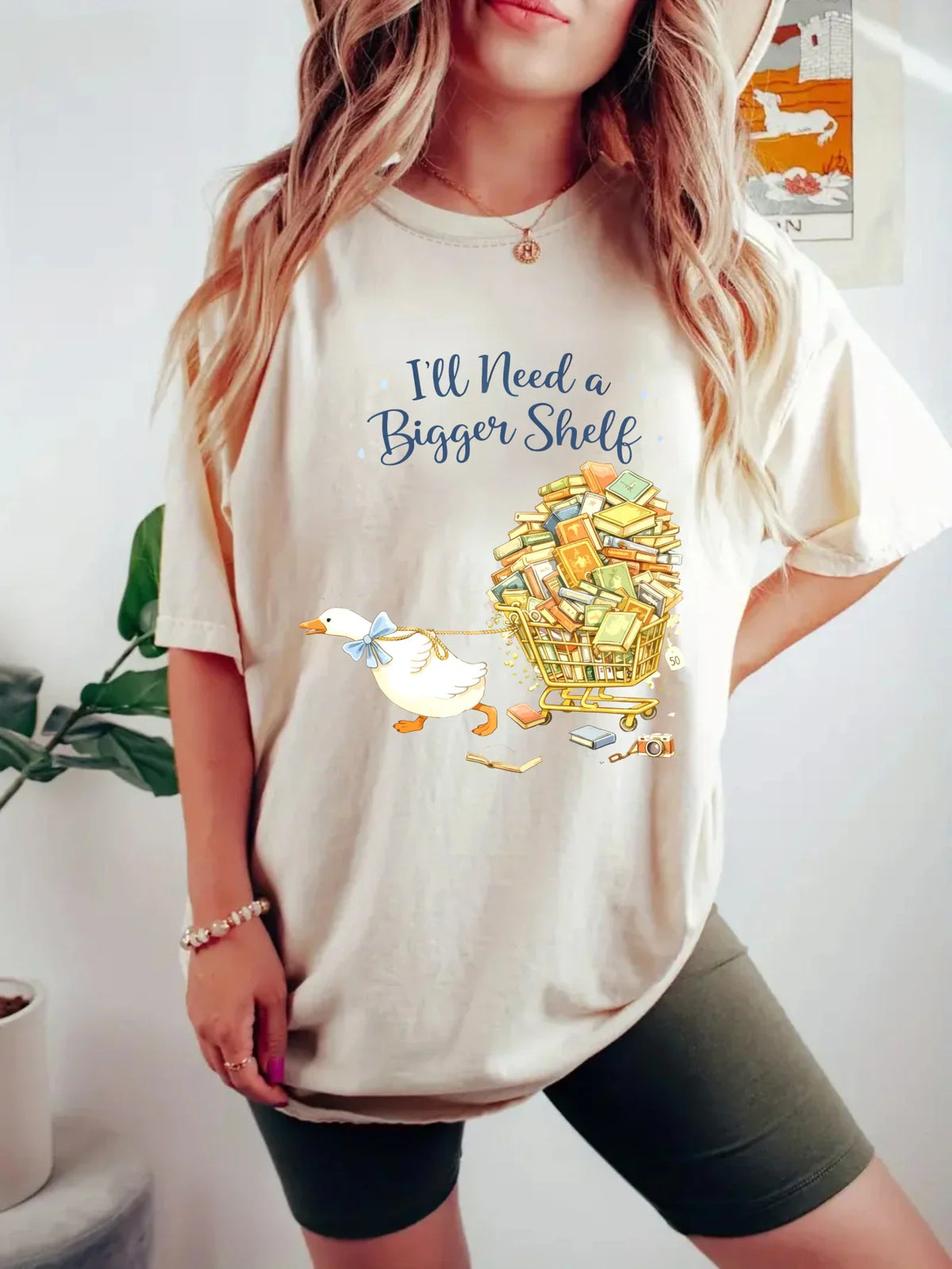 Beige short-sleeved t-shirt with a duck pulling a book cart graphic print, worn by a woman with green shorts