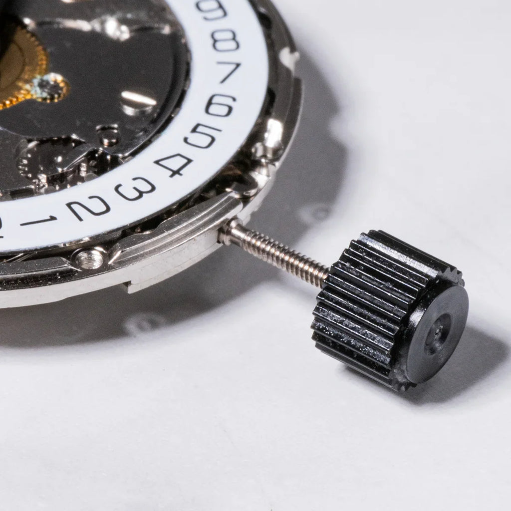 A close-up of a silver mechanical watch movement with a date wheel and a black winding stem.