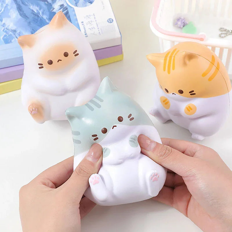 A hand squeezing a grey and white cat stress relief toy with other toy cats and books in the background.